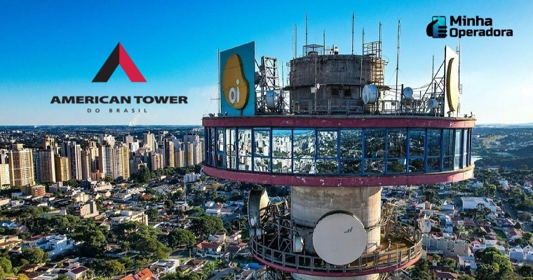 A imagem mostra uma torre de telecomunicações da Oi, destacada contra o céu azul. A parte superior da estrutura é circular, com janelas de vidro ao redor, refletindo o ambiente ao redor. No topo da torre, várias antenas e equipamentos de telecomunicações são visíveis. O logo da Oi aparece em dois grandes painéis na lateral da estrutura. Ao fundo, há uma vista panorâmica de uma área urbana, com prédios residenciais e comerciais cercados por árvores. Essa torre provavelmente faz parte da infraestrutura que a Oi está vendendo como parte de seu processo de reestruturação financeira.